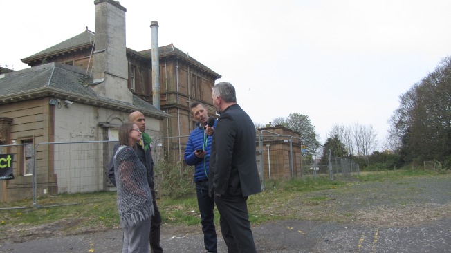 Photograph of interviewer with microphone interviewing 3 people in front of derelict building